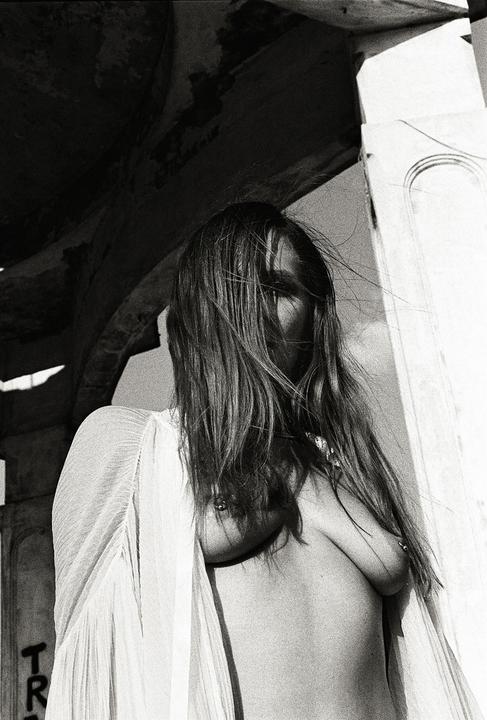 A woman with long hair covering her face stands topless in a sheer top.