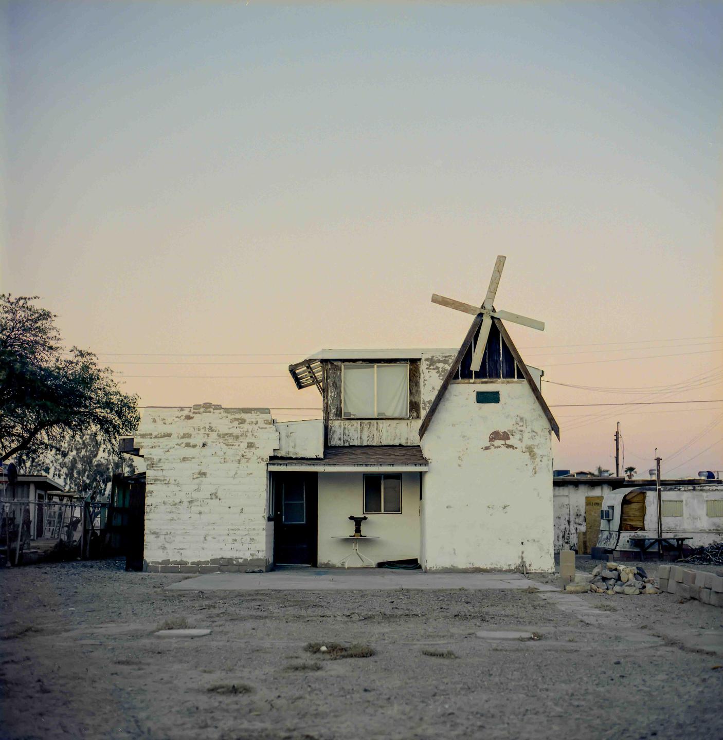 A white building with a windmill on its roof stands in a dusty lot under a pale sky.