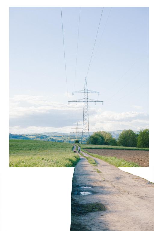 A person walks a dog down a dirt path next to a field with a large power line tower in the distance.
