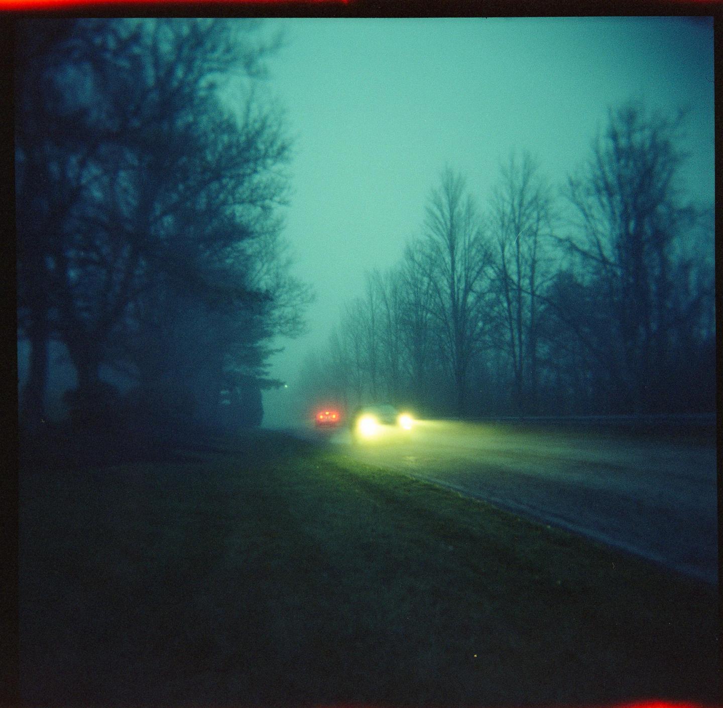 A car with bright headlights and red taillights drives down a road lined with trees in hazy conditions.