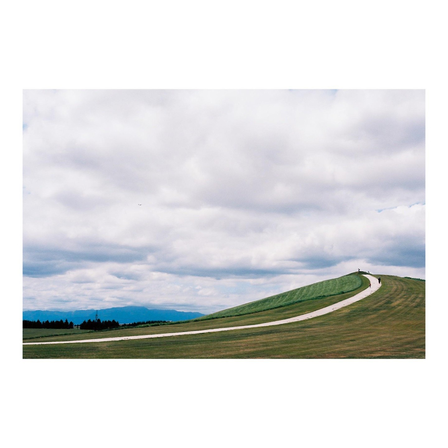 A winding path leads up a grassy hill under a cloudy sky, with a small airplane visible in the distance.