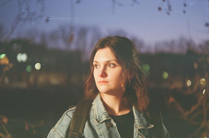 A woman with a denim jacket and a backpack looks to the side at dusk.