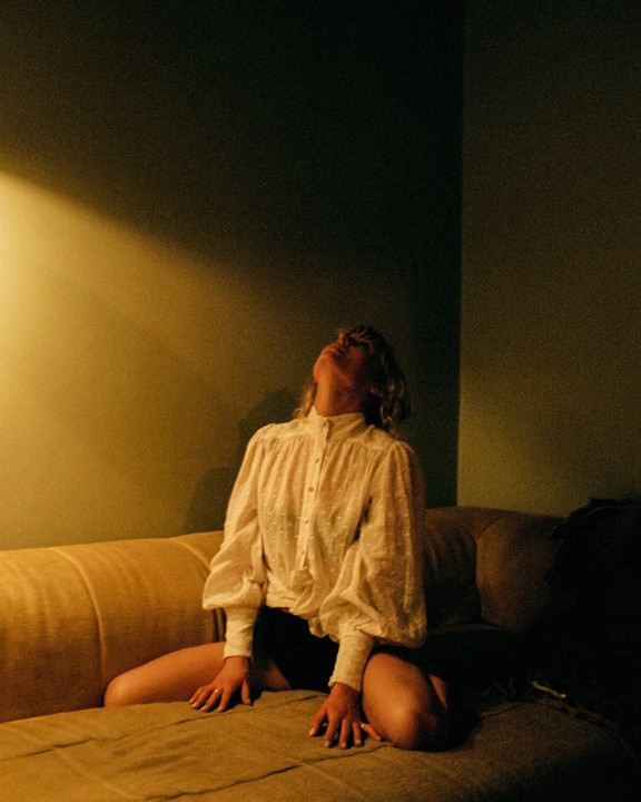 A woman in a white blouse and dark shorts kneels on a sofa, looking up at a beam of light.