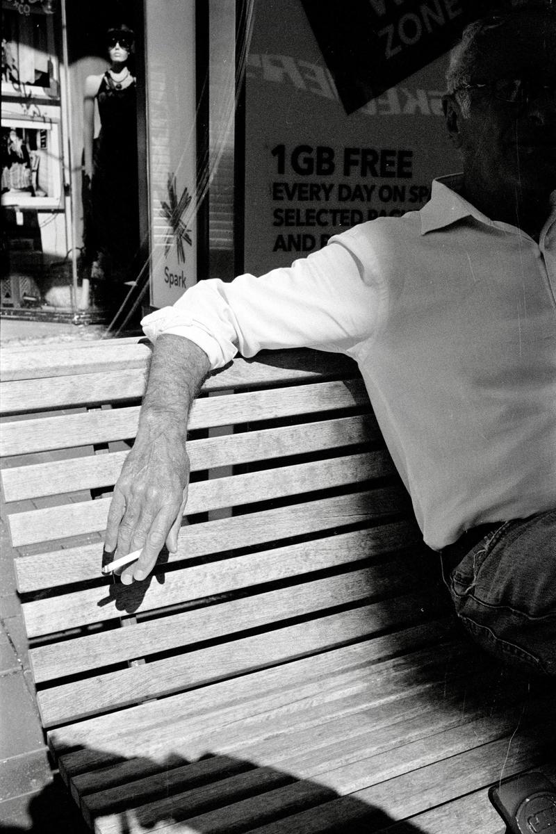 A man sits on a wooden bench with a cigarette in his hand, with a store window and mannequin visible in the background.