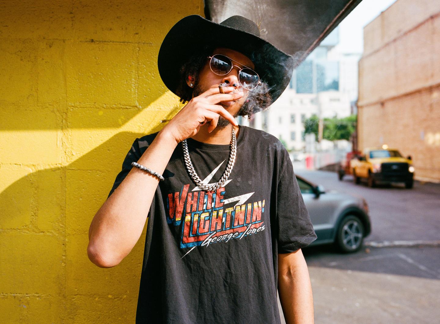 A man wearing a hat and sunglasses smokes a cigarette on a street next to a yellow wall.