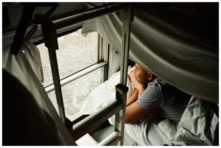 A man lies in a bunk bed on a train, looking out the window.