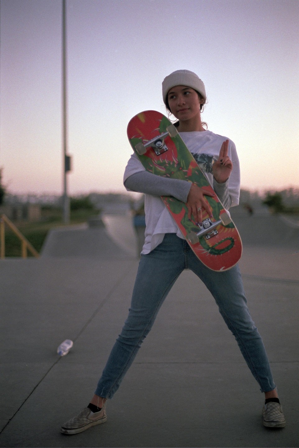 A young woman wearing a beanie and jeans holds a skateboard at a skate park during dusk.