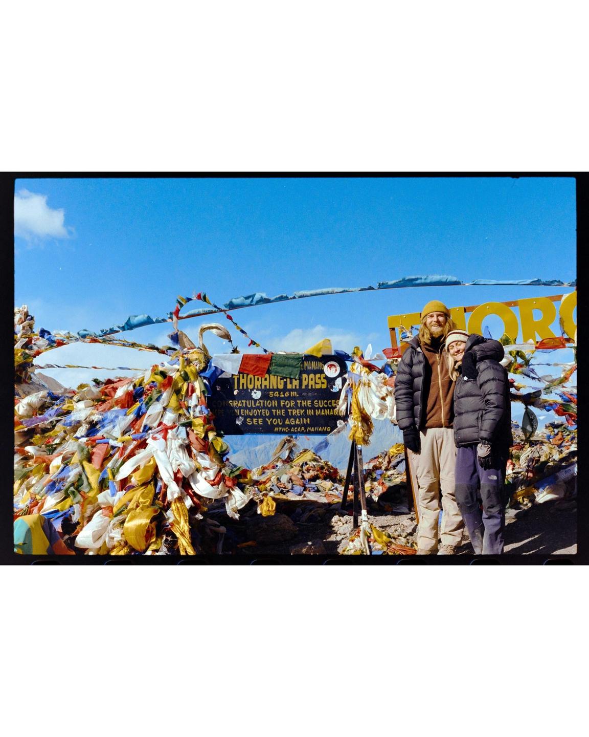 A couple stands in front of a sign and many colorful prayer flags on a mountain.