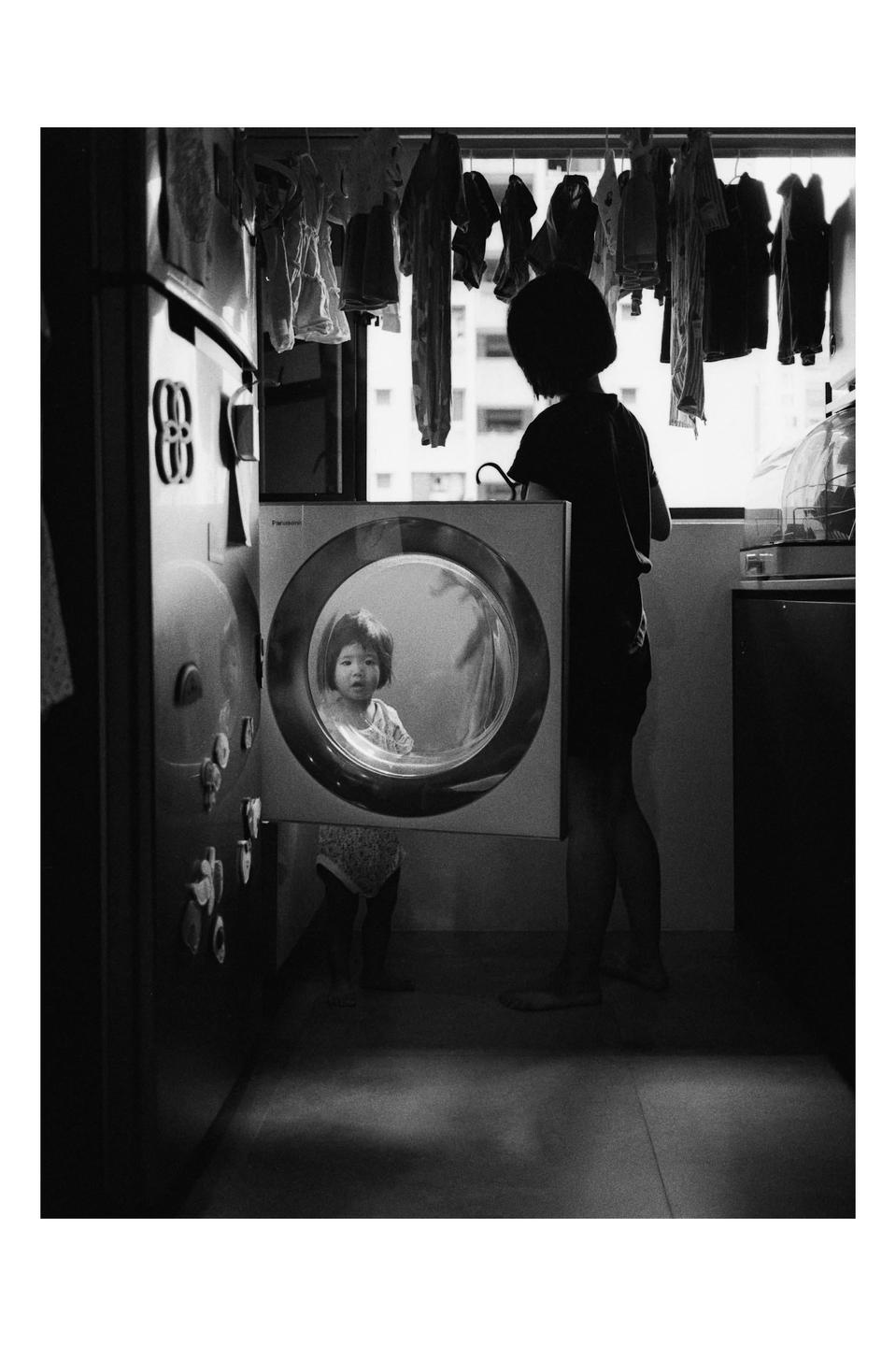 A child looks into a washing machine while another child stands with their back to the viewer in a room with clothes hanging to dry.