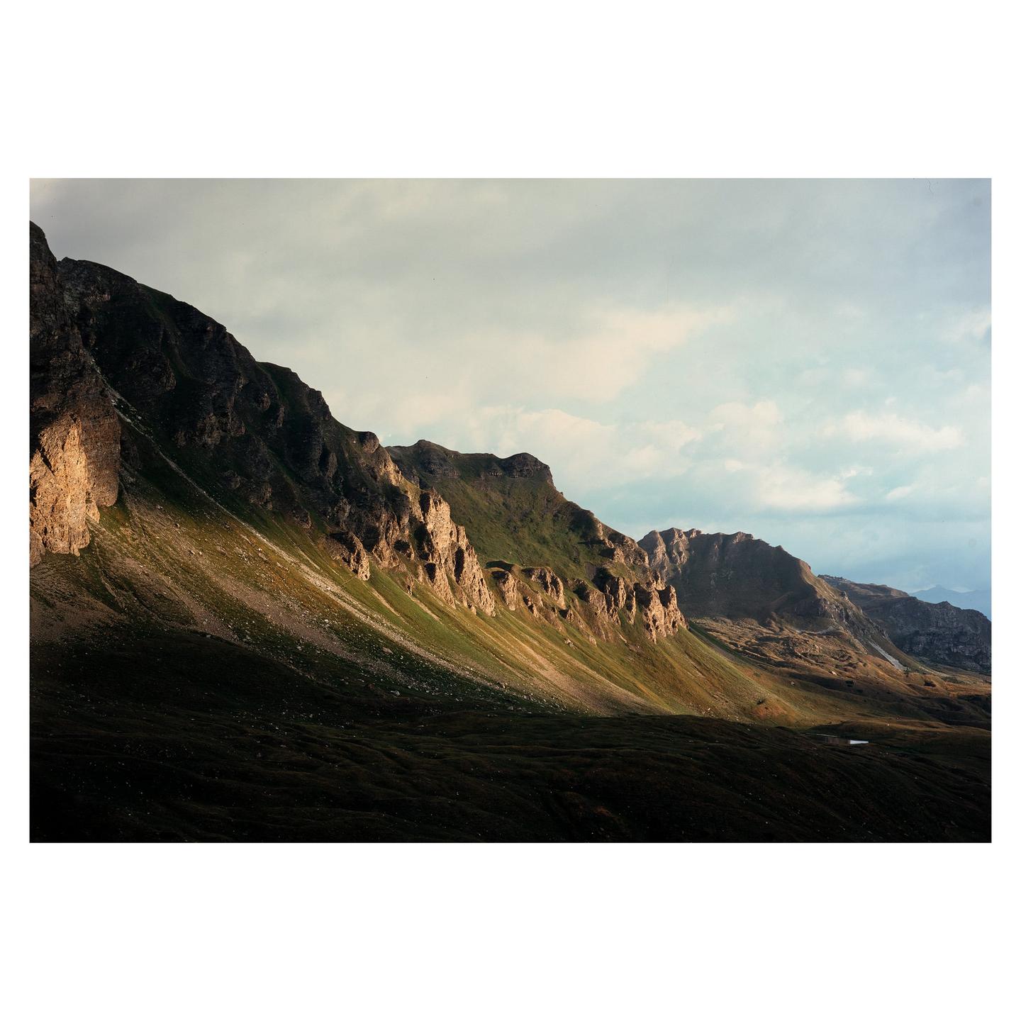 A dramatic landscape shows a mountain range with a valley in shadow and sunlit peaks under a cloudy sky.