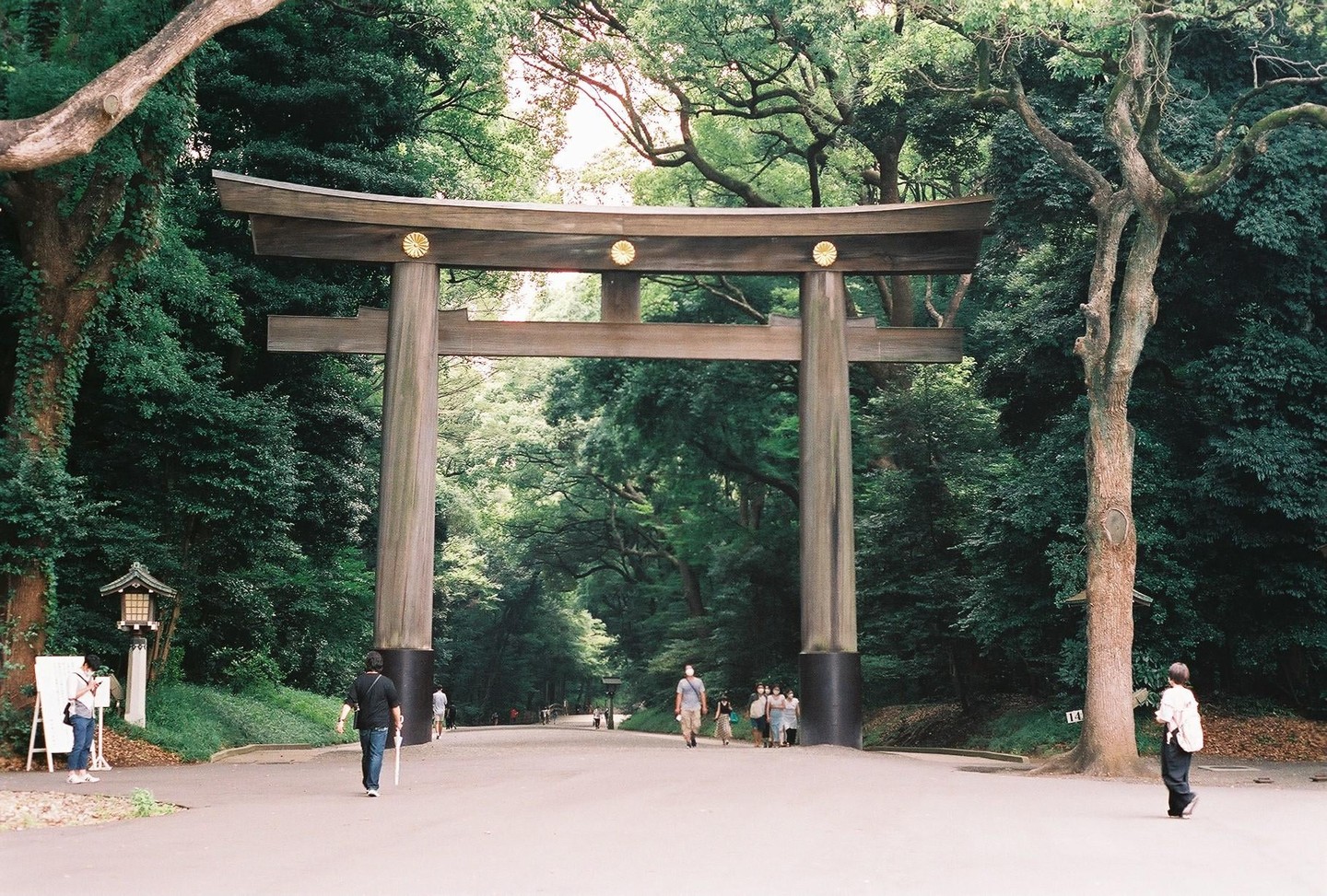 A large wooden torii gate stands at the entrance to a park, surrounded by lush green trees and several people walking on a paved path.