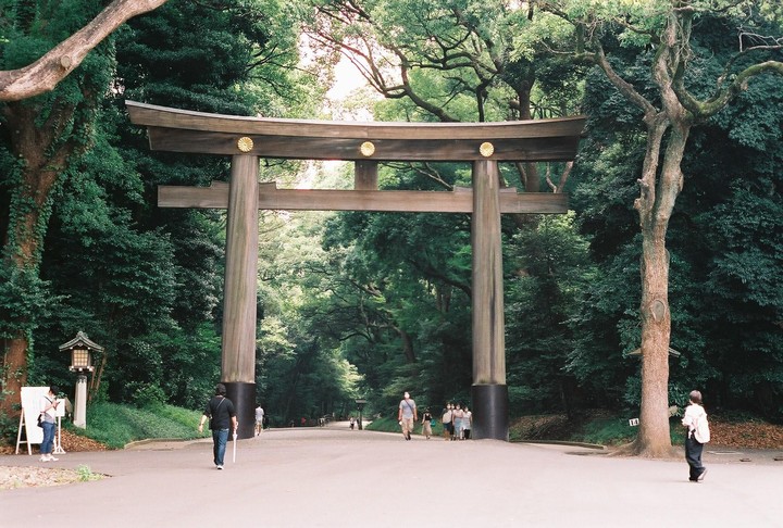 A large wooden torii gate stands at the entrance to a park, surrounded by lush green trees and several people walking on a paved path.