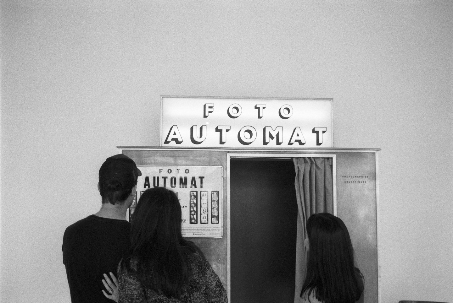 Three people stand in front of a photo booth with a sign that reads "FOTO AUTOMAT" in black and white.