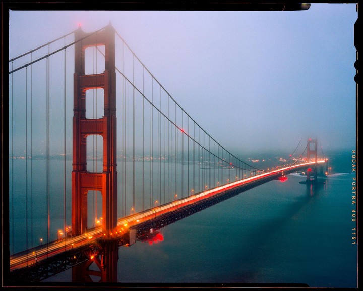 The Golden Gate Bridge is illuminated at dusk, with car light trails visible on the road.