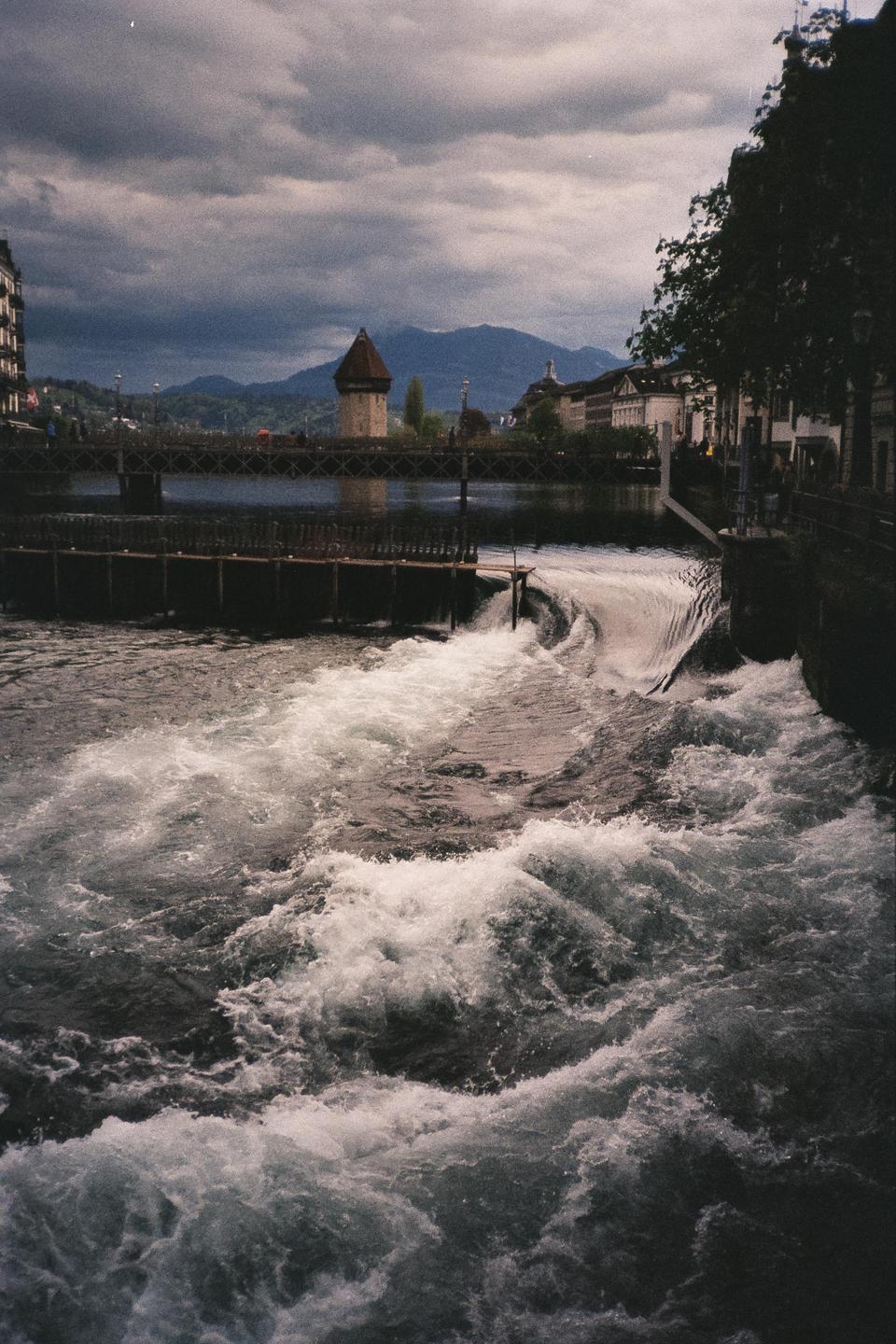 A river with rapids flows past a bridge and buildings under a cloudy sky.