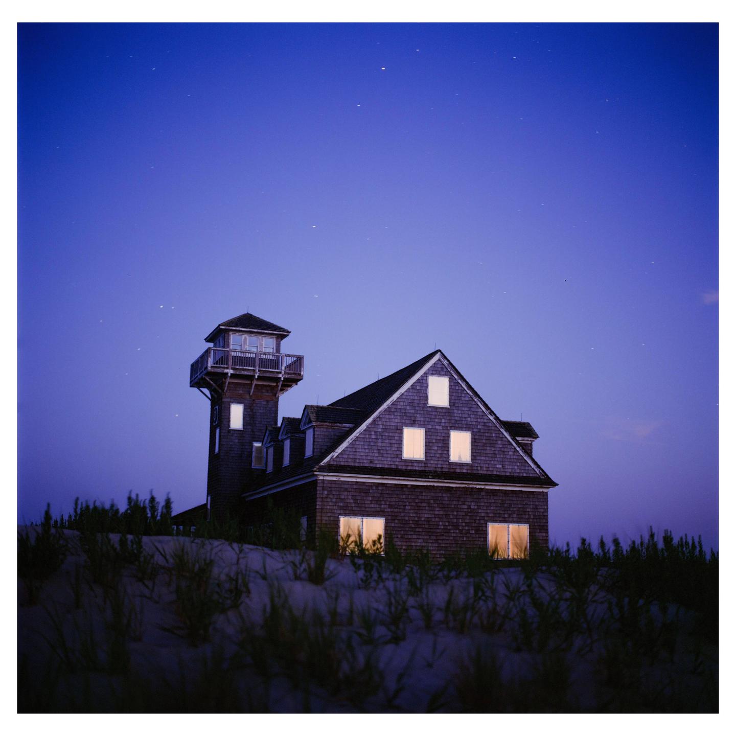 A house with a tower and illuminated windows stands on a sandy dune under a starry night sky.