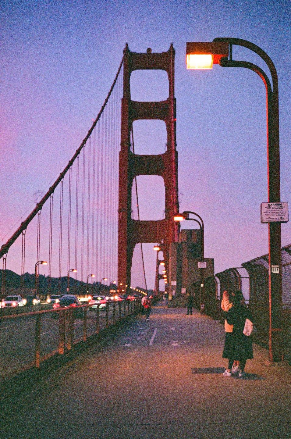 A couple stands on the Golden Gate Bridge at dusk, with cars driving past and the city lights in the distance.