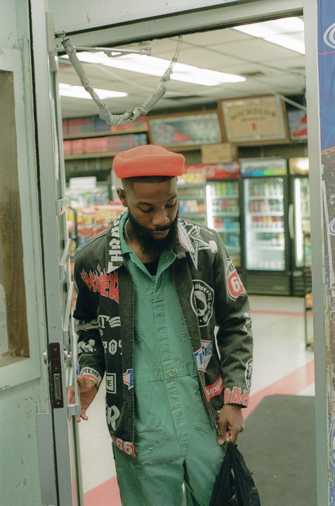 A man in a green jumpsuit and a patched jacket stands in a doorway, holding a black bag.