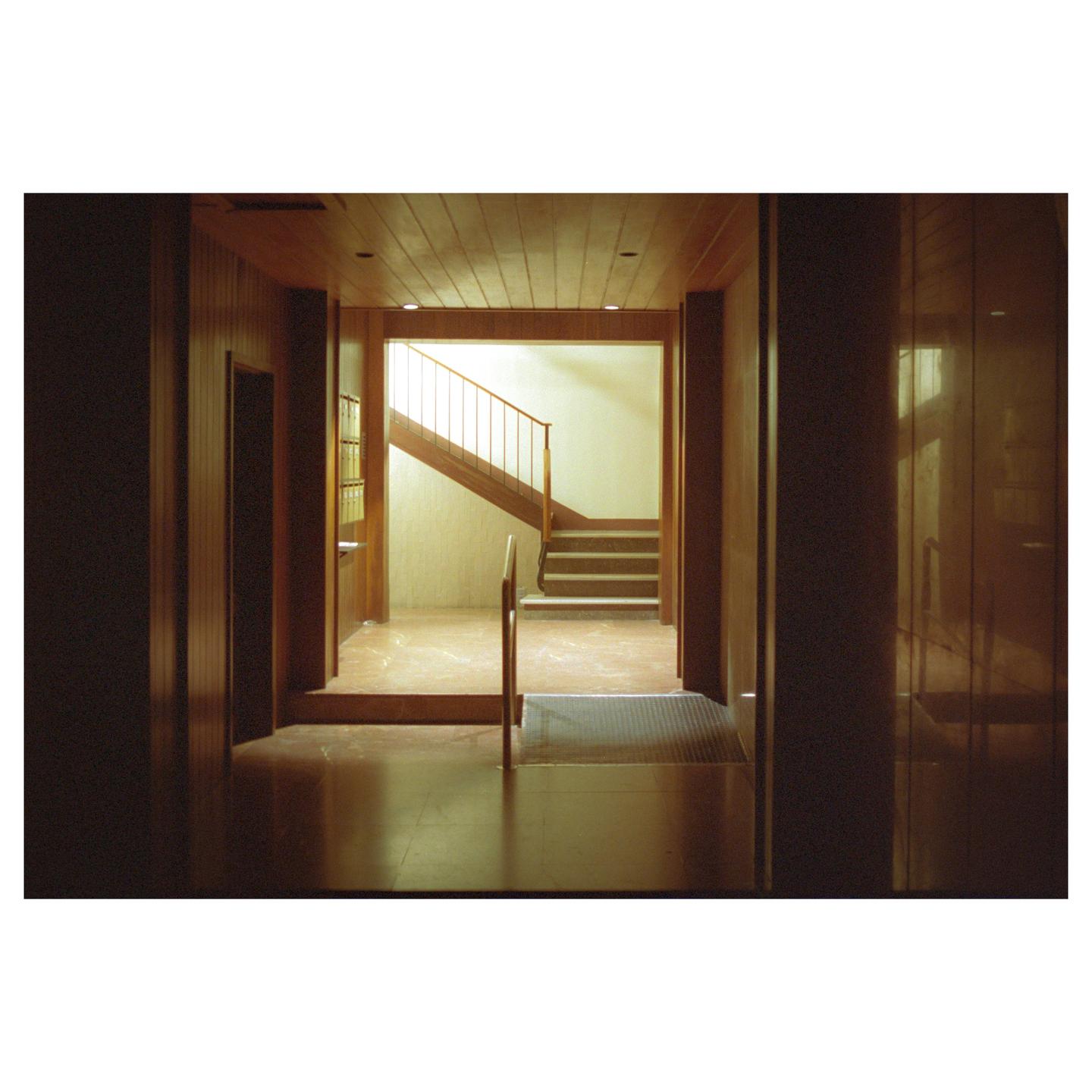 A wooden hallway leads to a brightly lit stairwell with a metal railing.