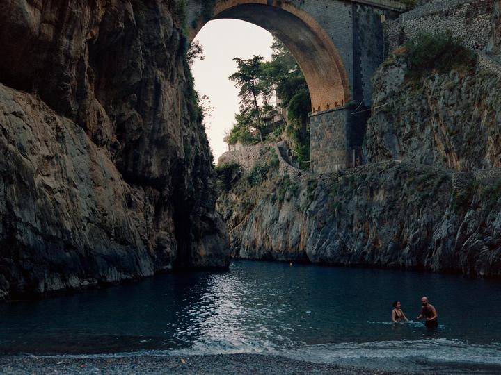 A couple wades in the water under a large stone bridge and rocky cliffs.