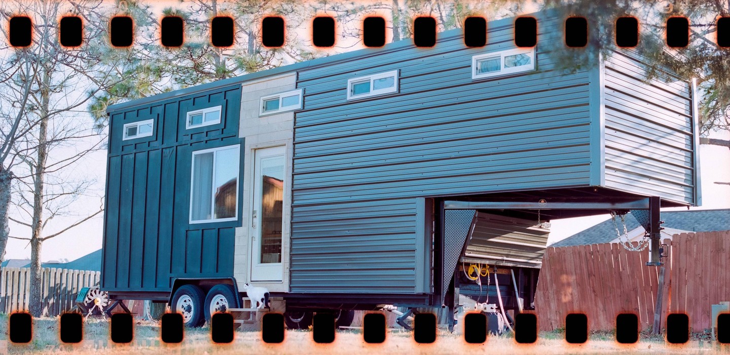A tiny house on wheels with a cat on the steps is parked in a yard with a wooden fence.