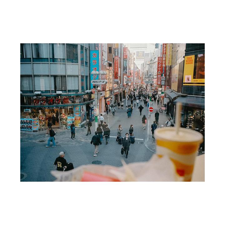 A busy street in Shibuya, Tokyo, is filled with pedestrians and neon signs, with a drink in the foreground.