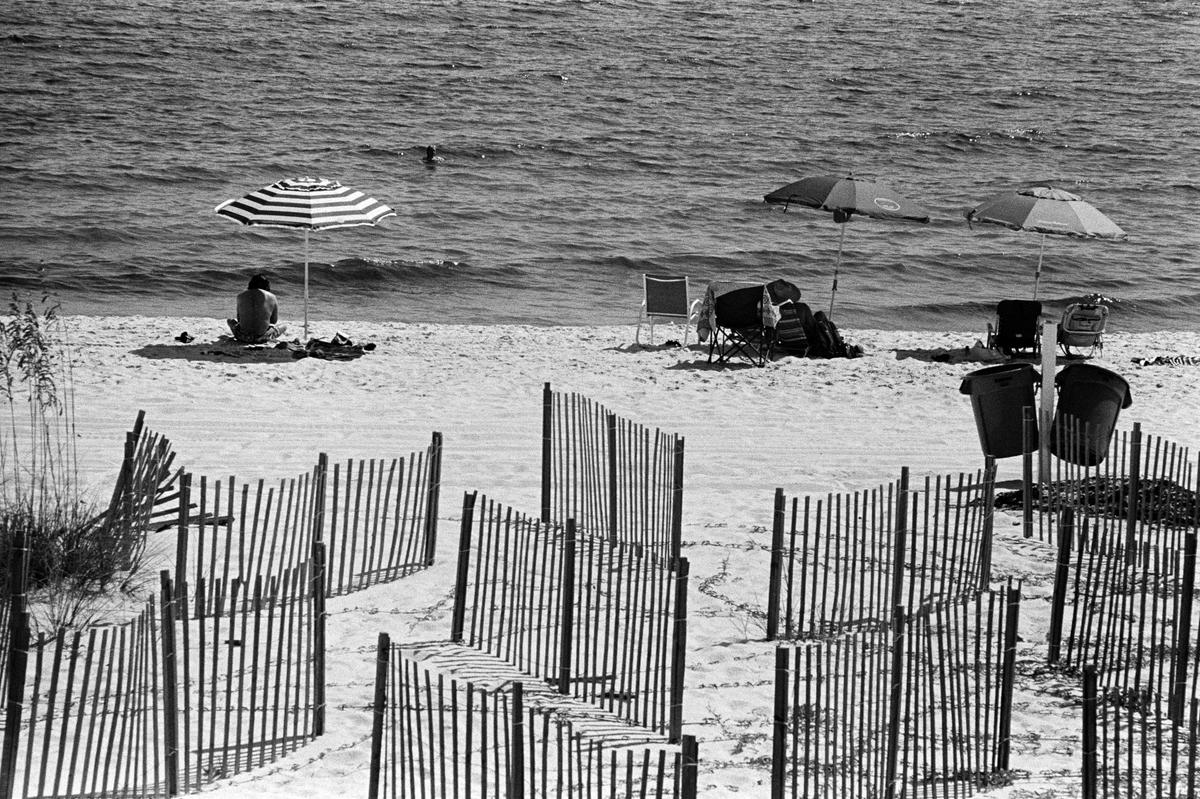 A person sits under a striped umbrella on a sandy beach with fences in the foreground and the ocean in the background.