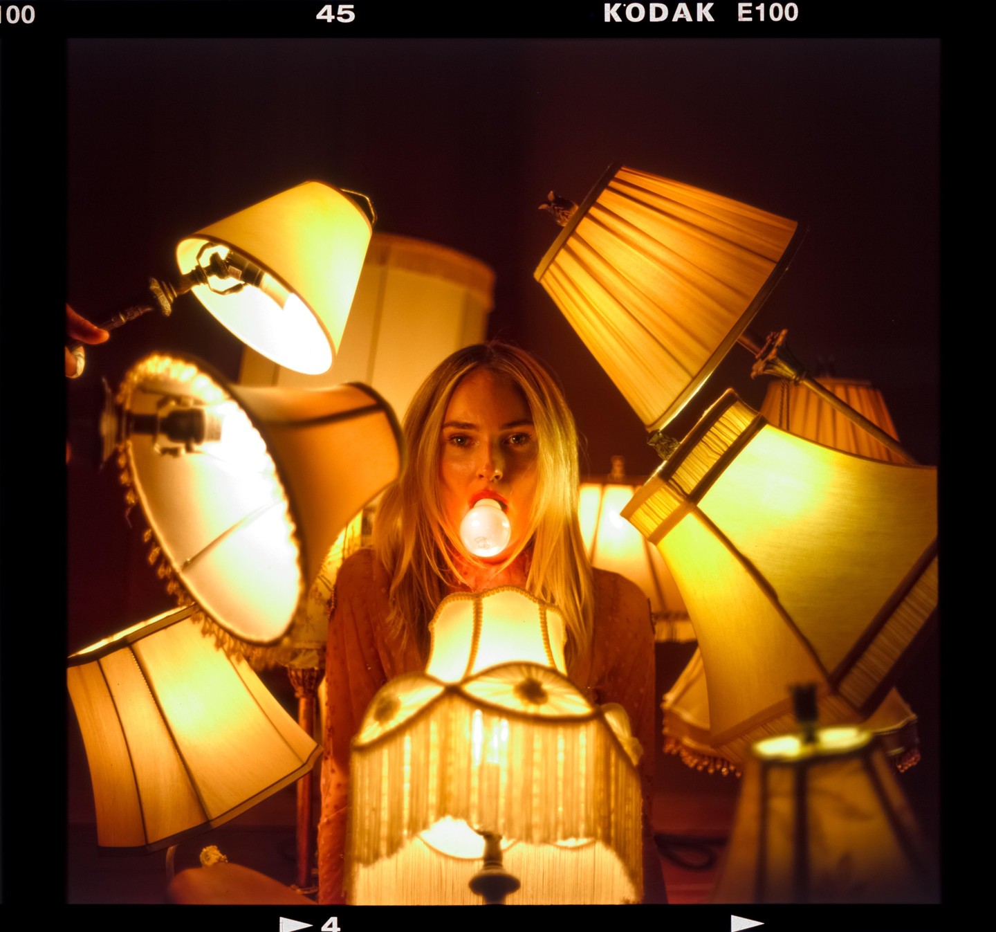 A woman with long hair holds a lightbulb in her mouth surrounded by many lamps.