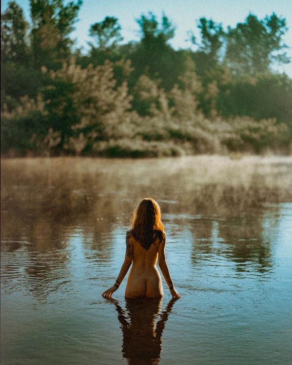 A woman with long red hair and tattoos on her back and arms stands in a body of water with trees in the background.