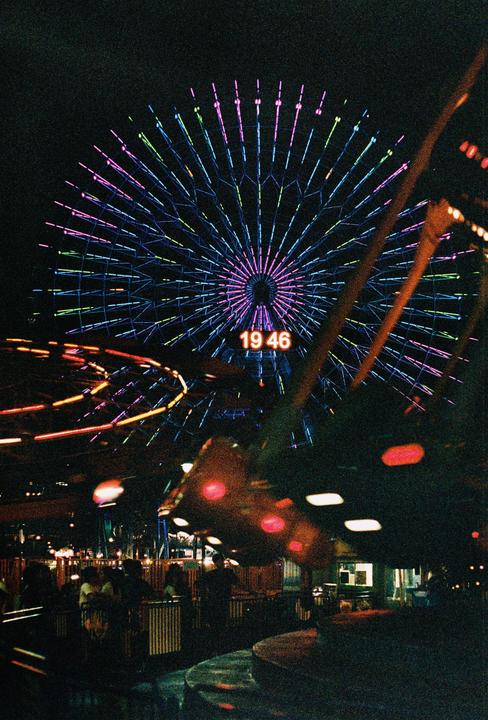 A ferris wheel with colorful lights and a digital clock displaying "19 46" is visible at night.