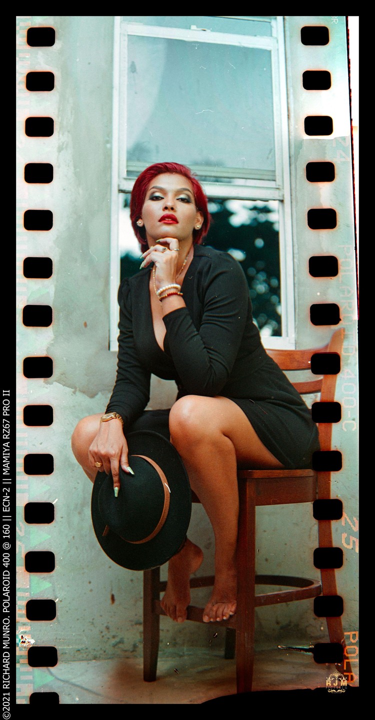 A woman with red hair and a black dress sits on a wooden chair, holding a hat.