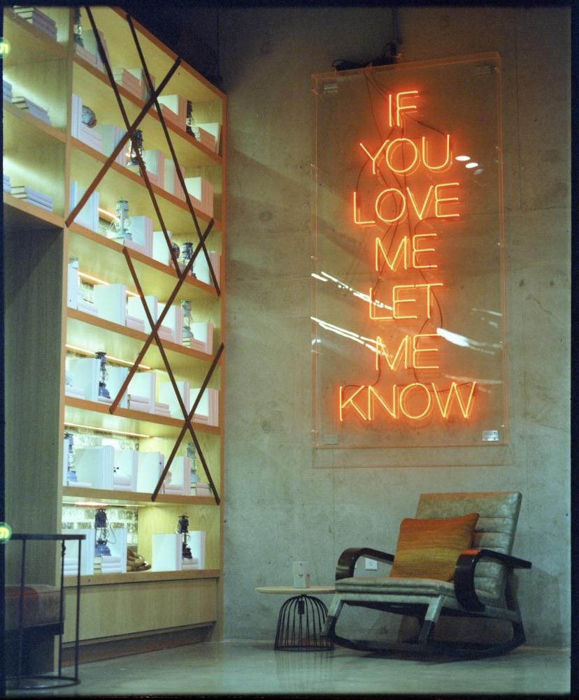 A neon sign with the words "IF YOU LOVE ME LET ME KNOW" hangs on a concrete wall next to a bookshelf and a rocking chair.