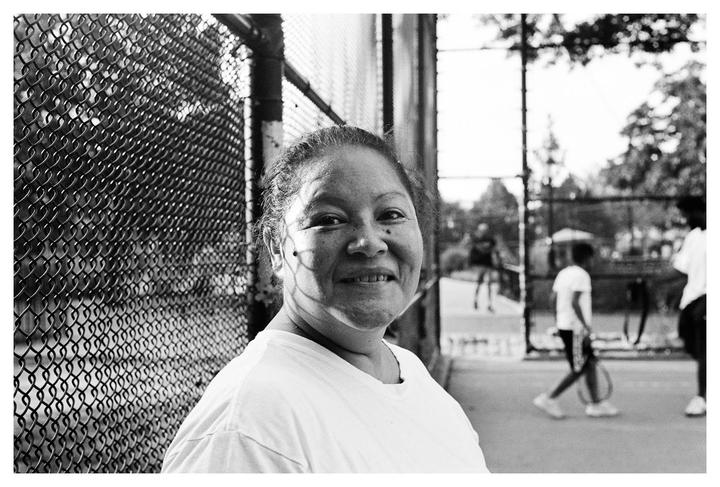 A woman smiles at the camera while standing next to a chain-link fence with people in the background.