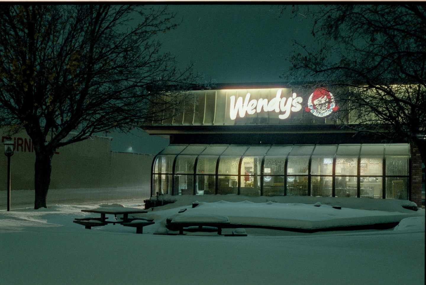 A Wendy's restaurant with a brightly lit sign is seen at night, surrounded by snow-covered ground and bare trees.