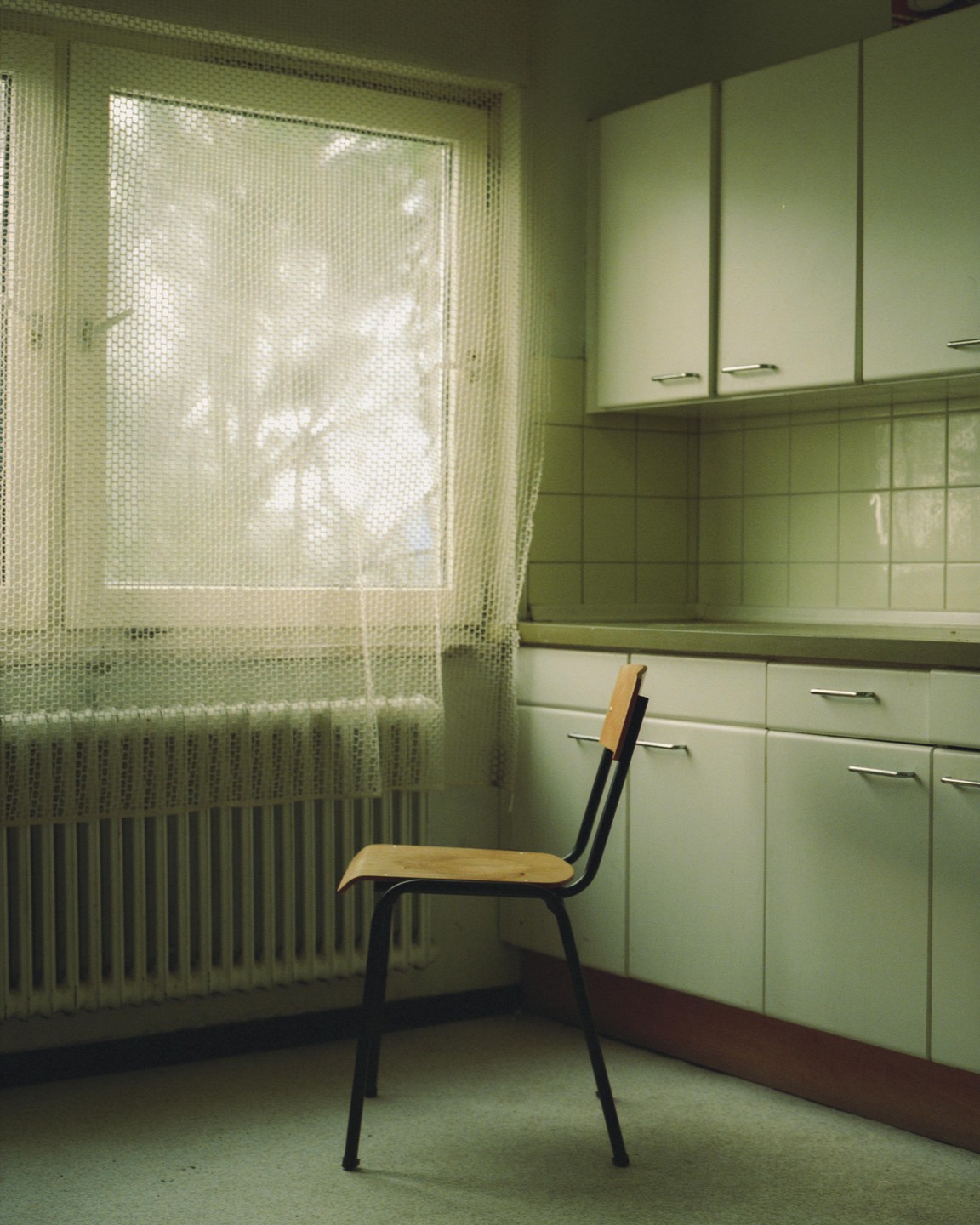 A wooden chair sits in a kitchen next to a window with a radiator underneath.