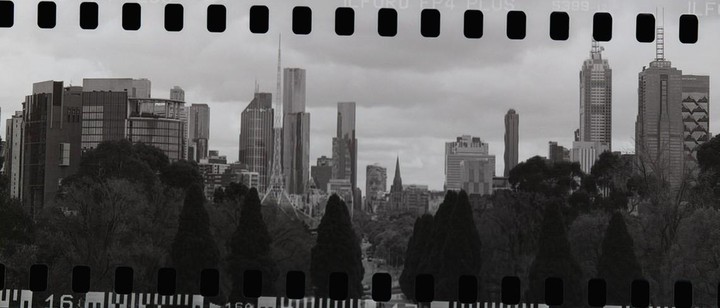 A panoramic view of a city skyline with tall buildings and a prominent spire, seen through a foreground of dark trees, with film sprockets visible at the top and bottom.