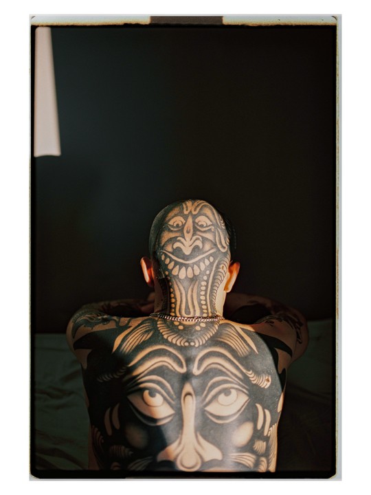 A man with extensive tattoos on his back and head is illuminated by a shaft of light.
