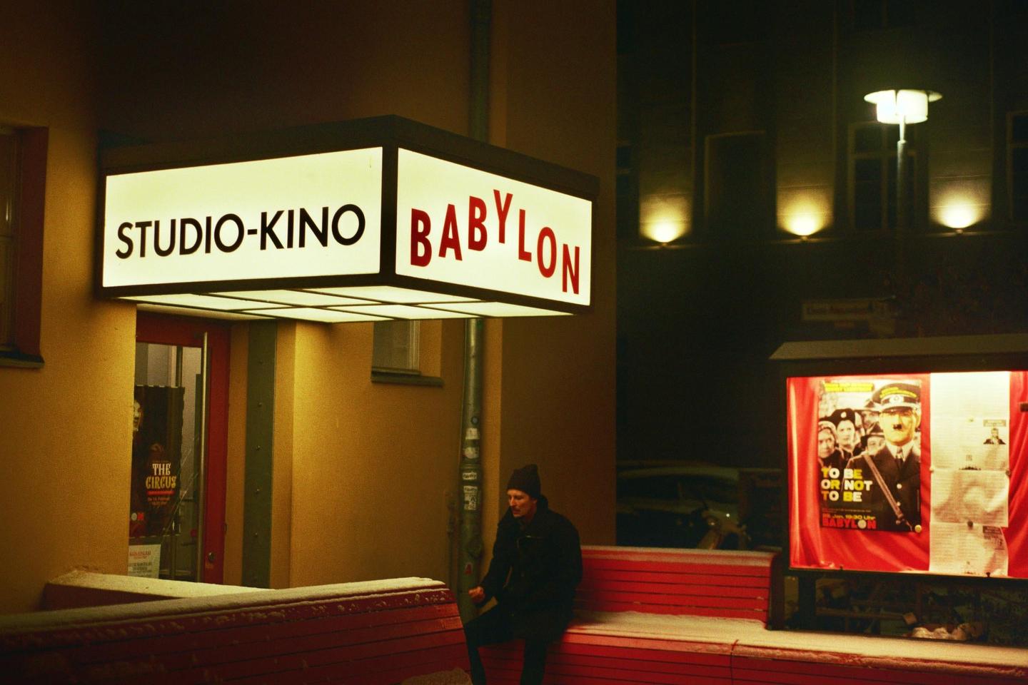 A man sits on a red bench outside the Studio-Kino Babylon at night, with snow on the ground.