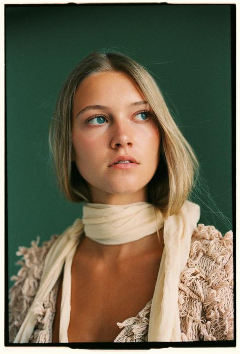 A woman with blonde hair and blue eyes wears a light scarf and a textured garment against a dark green background.