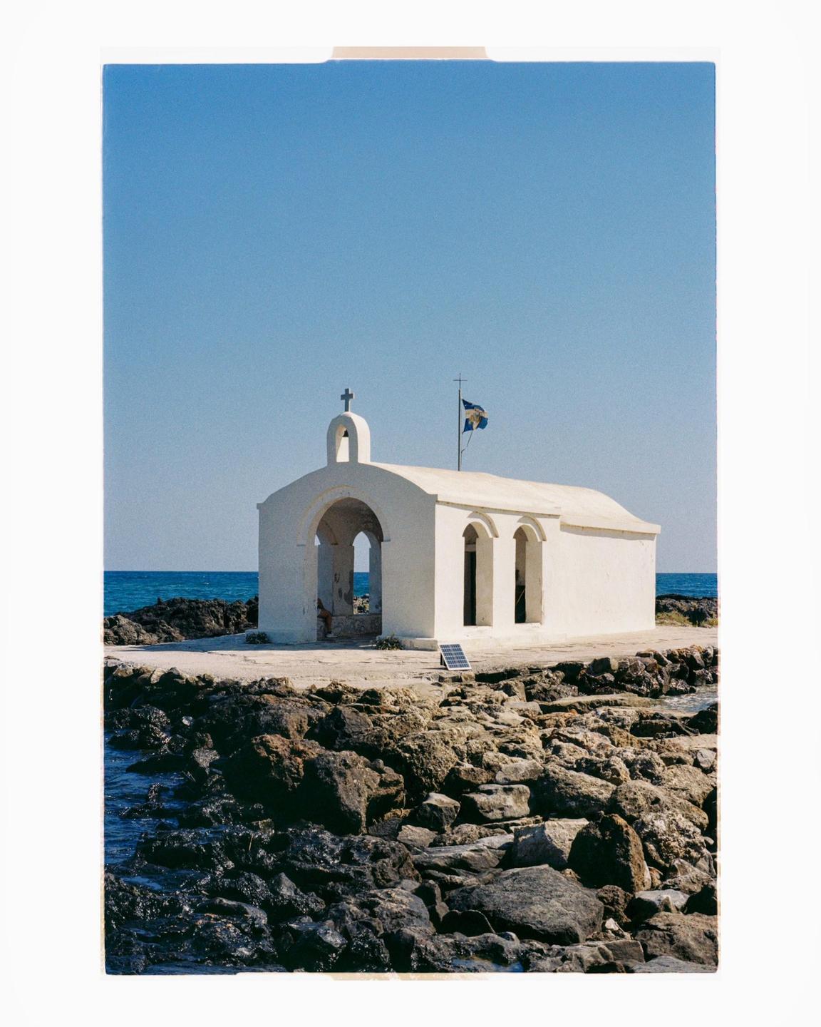 A small white church with a bell tower and a flag stands on a rocky shore next to the sea under a clear blue sky.