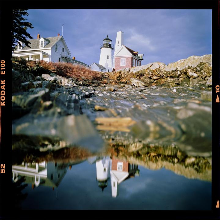 A white lighthouse and house are reflected in a tide pool on a rocky shore under a cloudy sky.