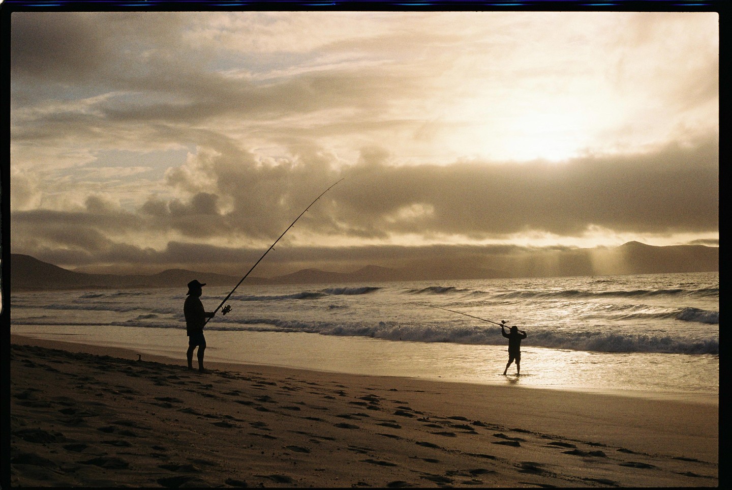A man and a boy are fishing on a beach at sunset with mountains in the distance.
