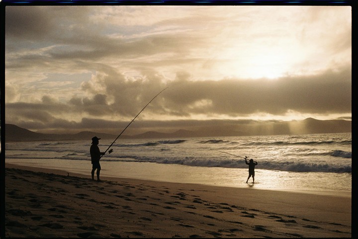 A man and a boy are fishing on a beach at sunset with mountains in the distance.