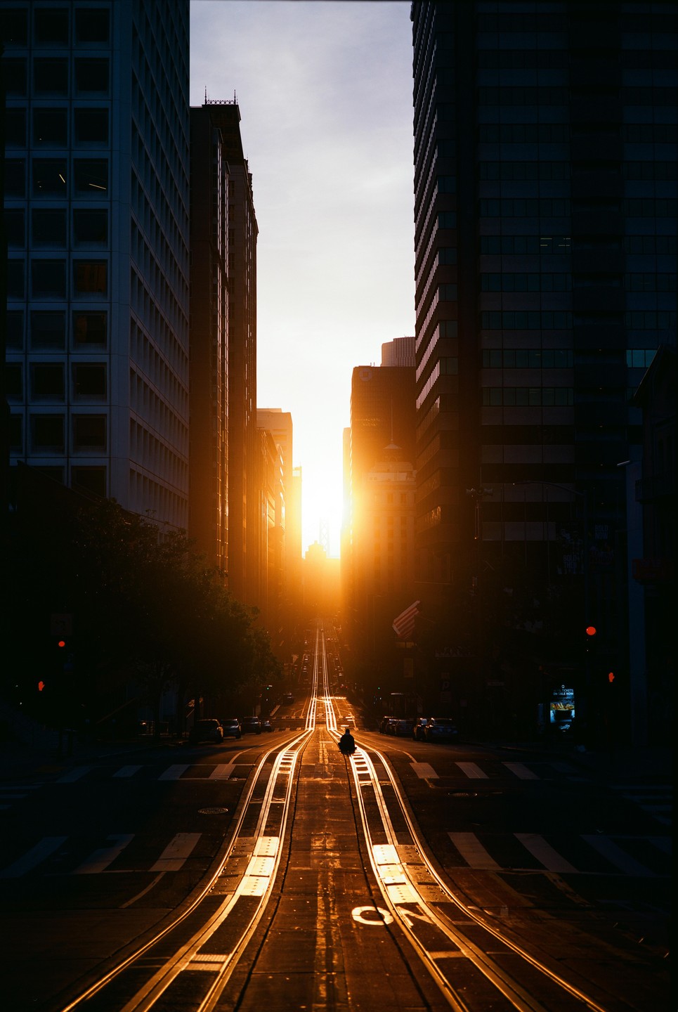 The sun sets at the end of a city street lined with tall buildings and cable car tracks.