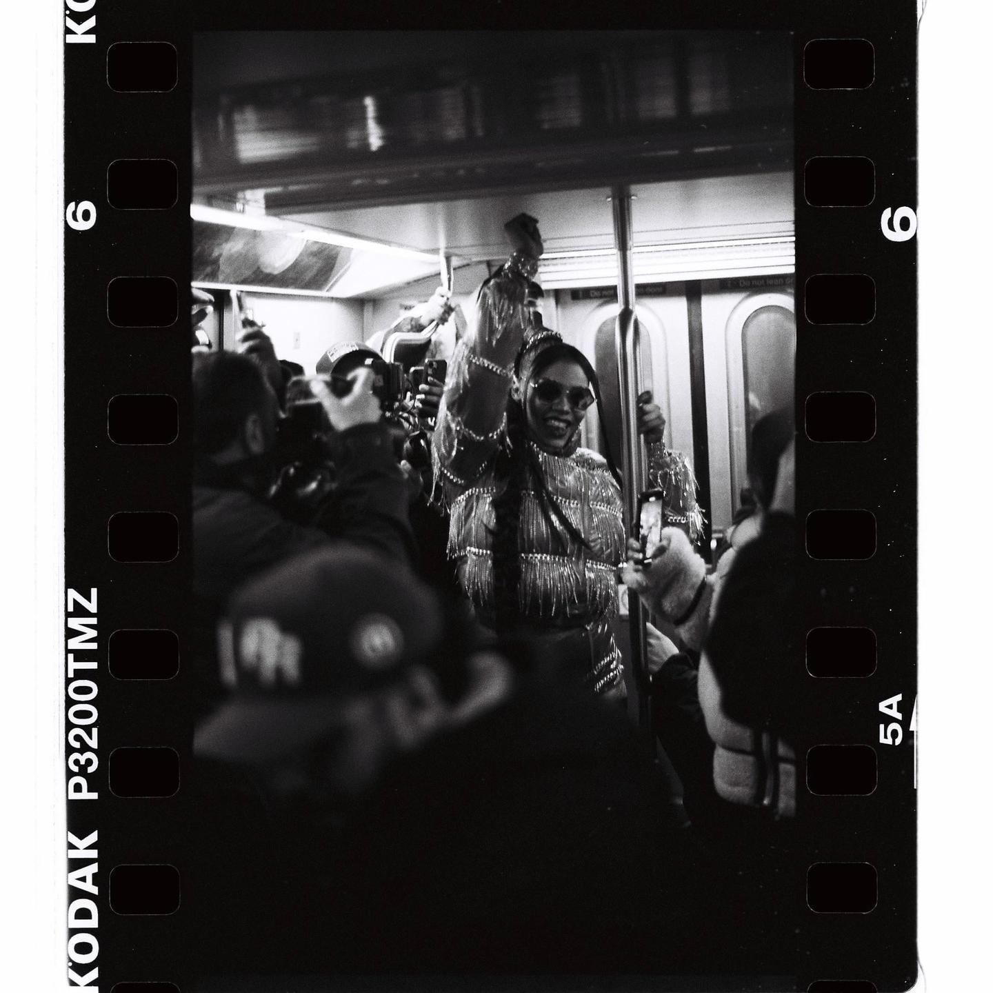 A woman in sunglasses and a fringed jacket smiles while holding onto a pole in a crowded subway car.