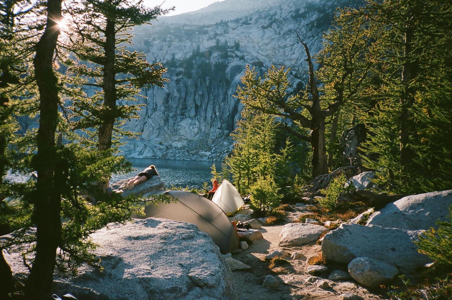 A woman lies on a rock near a lake with tents and trees in a mountainous landscape.