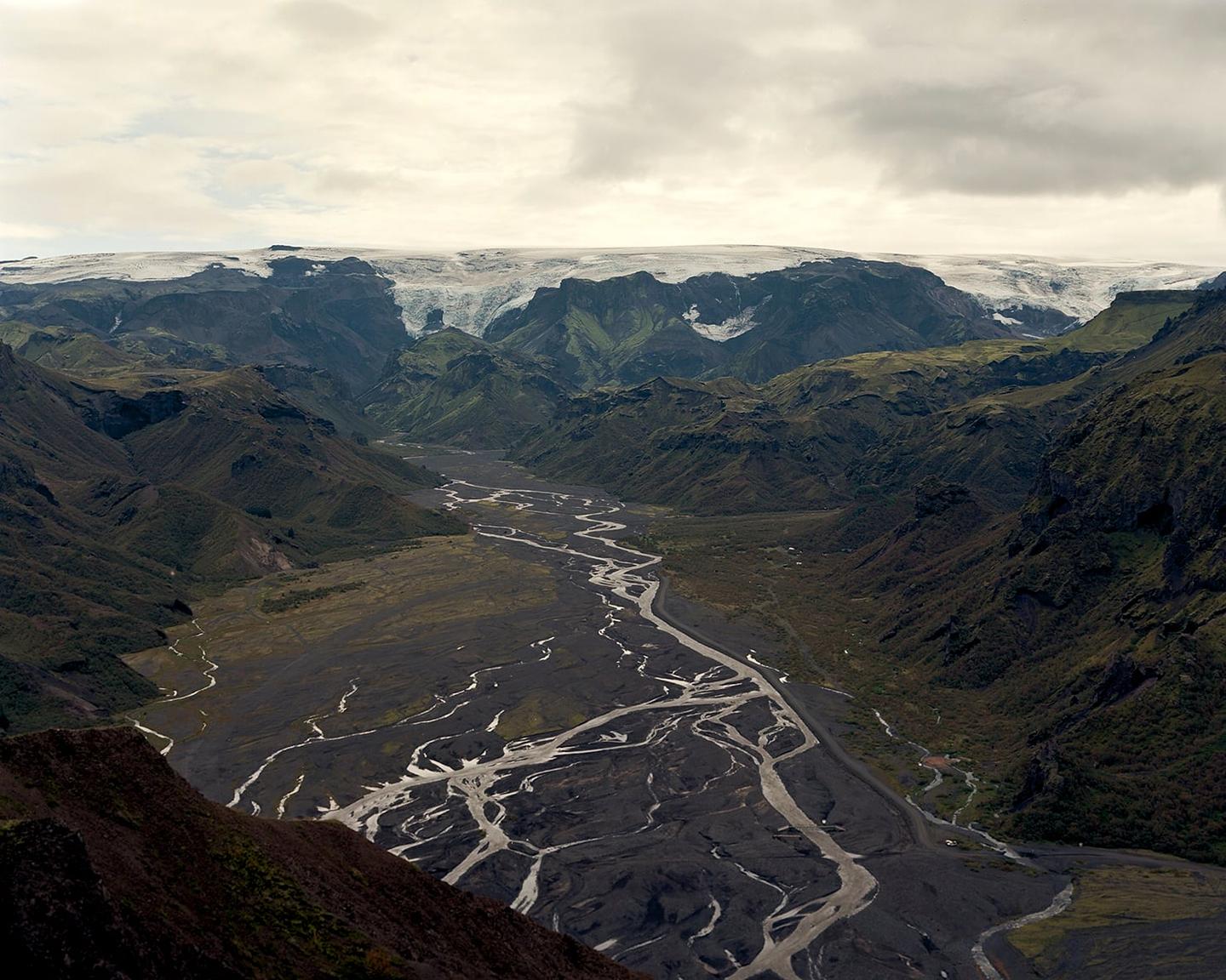 A wide river with many tributaries flows through a valley surrounded by mountains with snow-capped peaks under a cloudy sky.