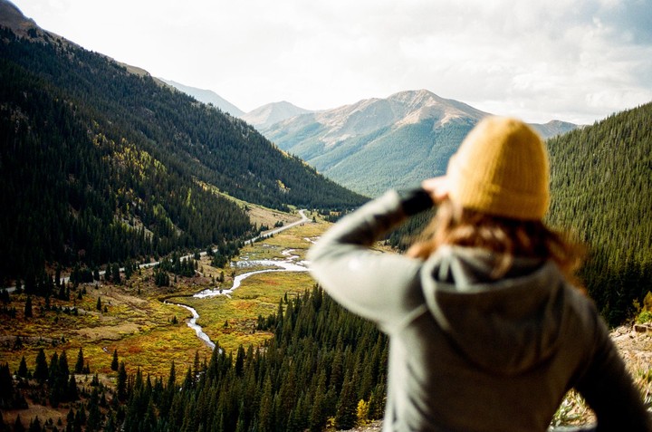 A woman in a yellow beanie looks out over a mountain valley with a winding river and road.