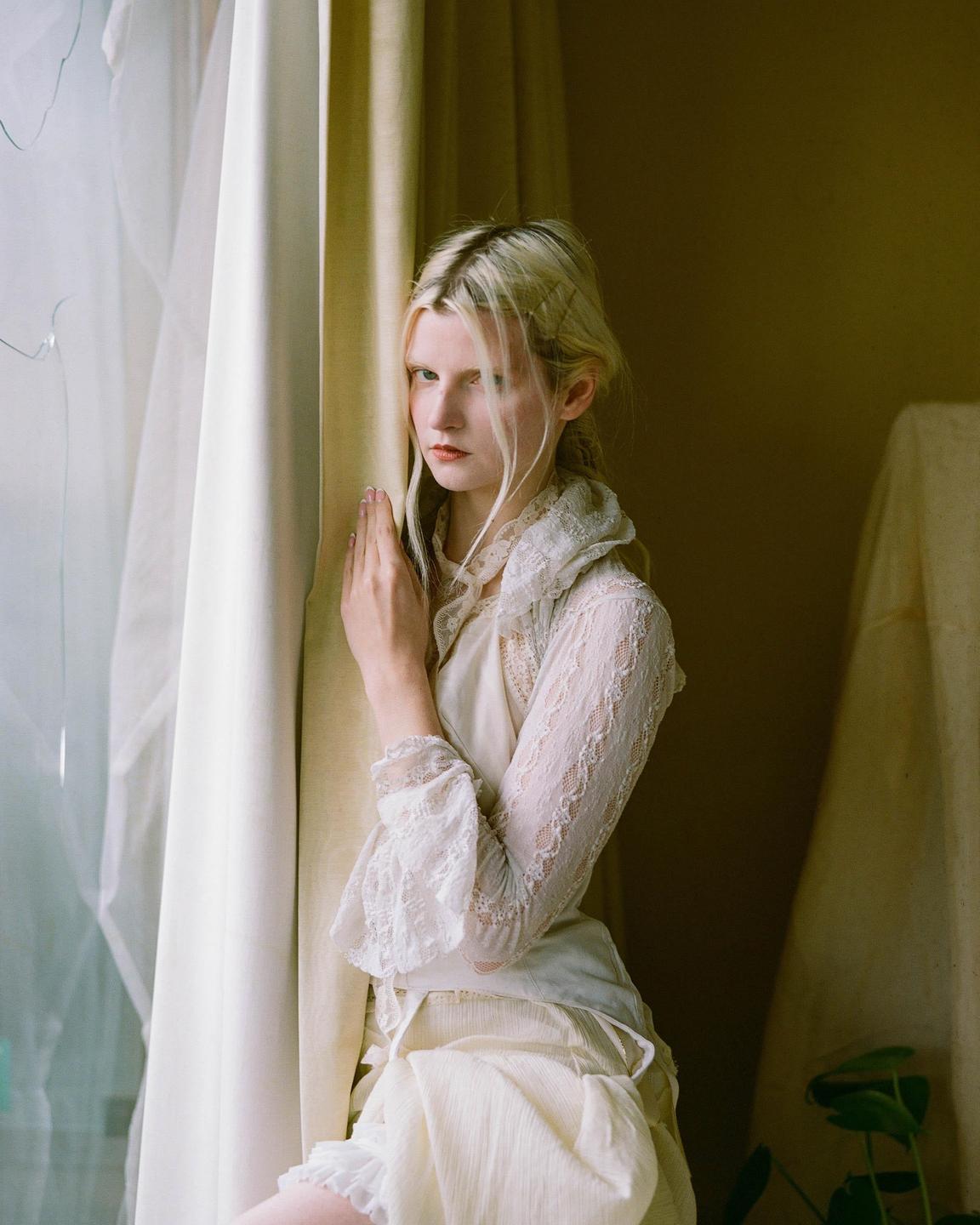 A woman with blonde hair and a white lace top stands by a window with curtains.
