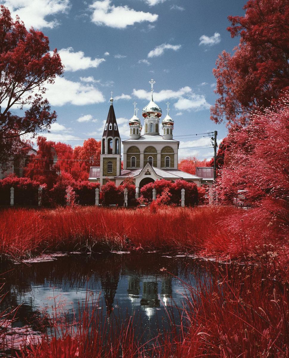 A Russian Orthodox church with silver domes is surrounded by red foliage and reflected in a pond.
